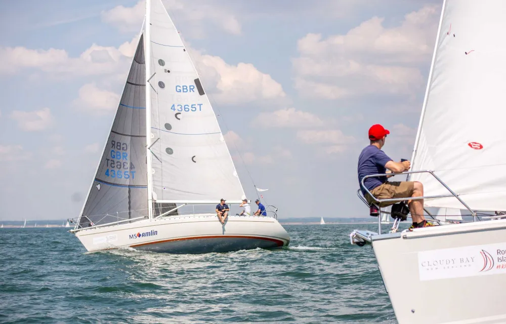 Two sailboats on a calm sea with sailors on board, one heading towards the other under a partly cloudy sky.