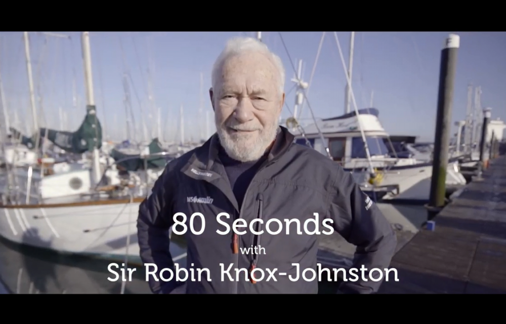 An elderly man stands in front of boats at a marina with text reading, 