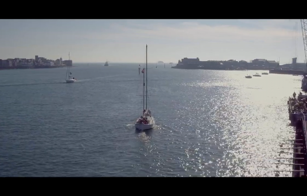 A sailboat navigates a calm, sunlit body of water with city buildings and other boats visible in the background.