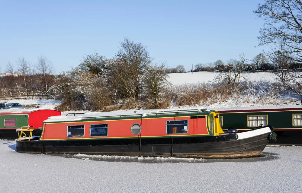 A narrowboat with a red and green exterior is moored on a frozen canal in a snowy landscape.