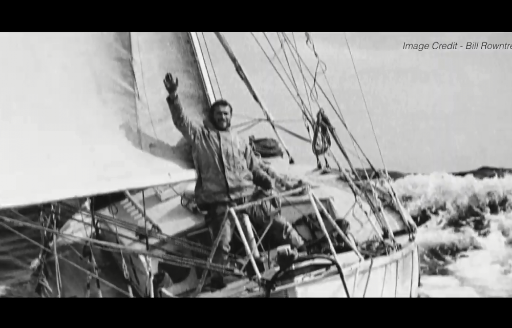 Person standing on a sailboat with raised hand in a greeting gesture, surrounded by ocean waves. Image credit: Bill Rowntree.