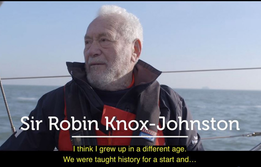 An older man in sailing gear stands on a boat; caption underneath reads, 