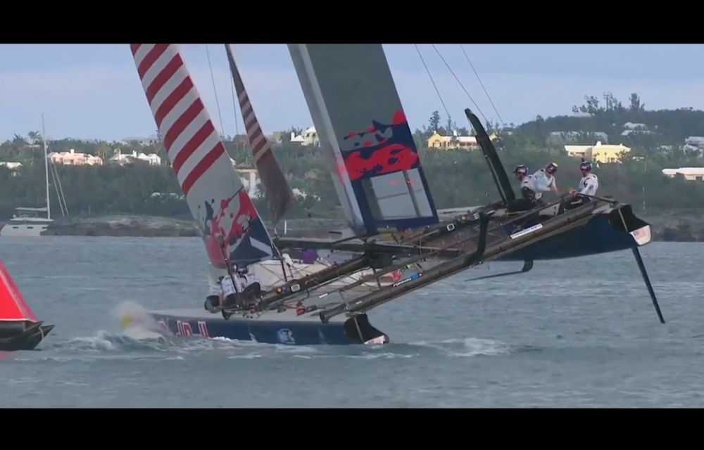 A catamaran, bearing colorful sails with a mix of red, white, blue, and stripes, is tilted on the water with crew members onboard.