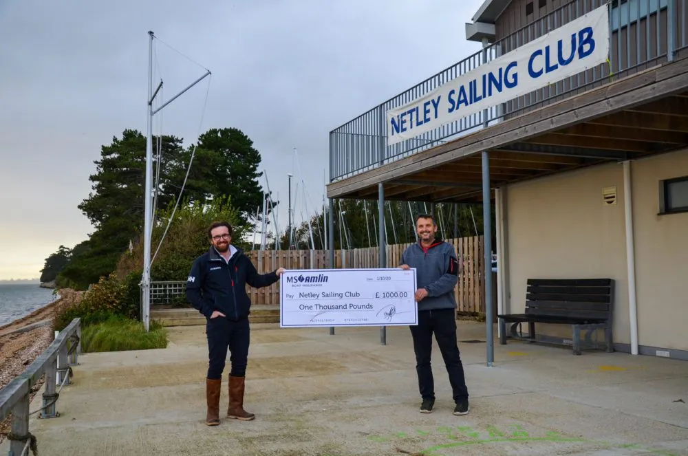 Two men standing outside Netley Sailing Club holding a large check for £1000. Sailboats and trees are in the background.