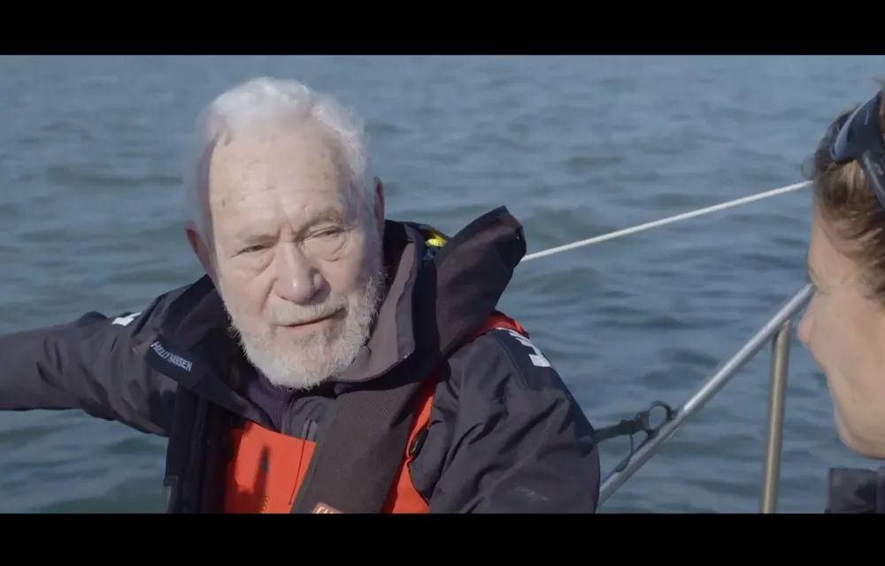 An elderly man with white hair and beard wearing a life jacket is sitting on a boat with water in the background.