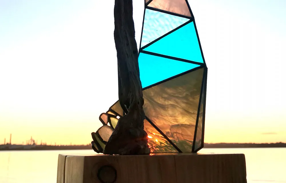 Stained glass sculpture with blue and green fragments set on a wooden base, with a sunset over water and industrial landscape in the background.