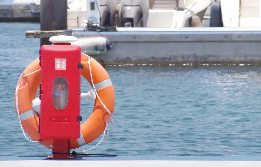 An orange lifebuoy mounted in a red holder by the water, with boats docked in the background.