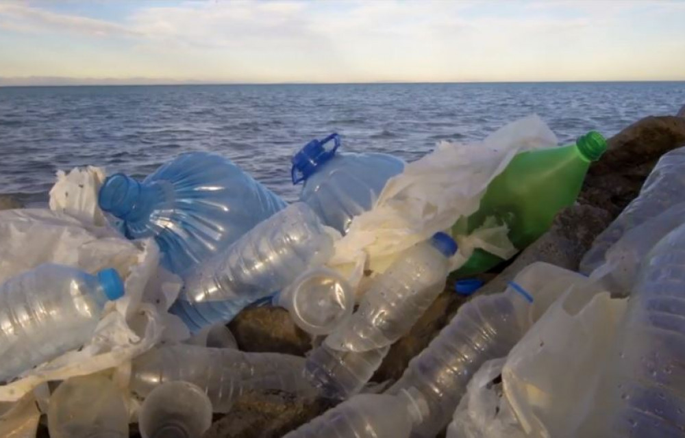 Plastic bottles and debris accumulated on a rocky shoreline with the ocean in the background.