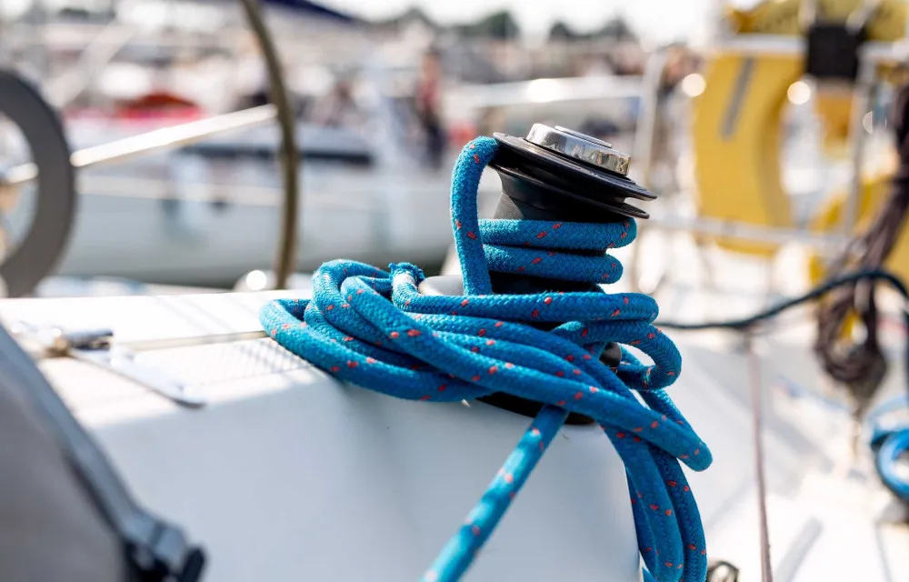 Close-up of a winch on a boat deck with blue rope coiled around it. Other boats and blurred background visible.