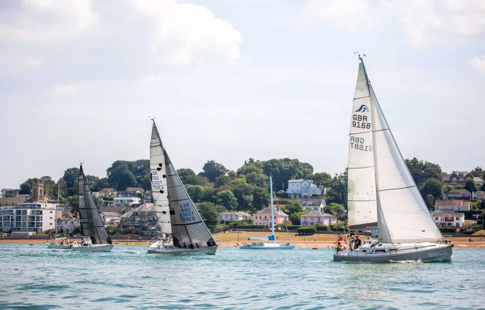 Several sailboats of varying sizes sail near a coastal town with buildings and trees in the background. The sky is partly cloudy.