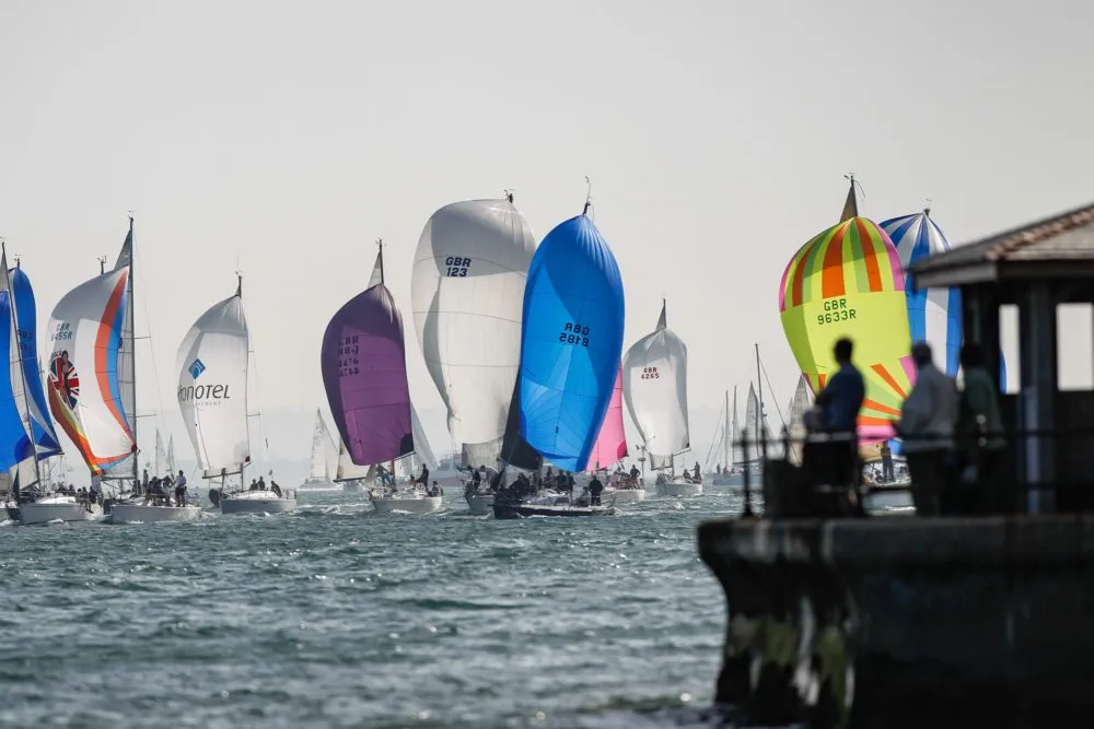 A fleet of colorful sailboats with spinnakers hoisted competes in a regatta on a sunny day, observed by people from a dock.