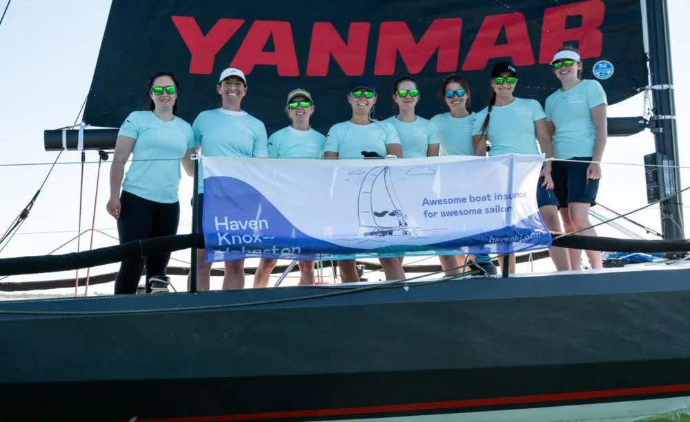 A group of nine people in matching light blue shirts stand on a boat, holding a banner that reads 