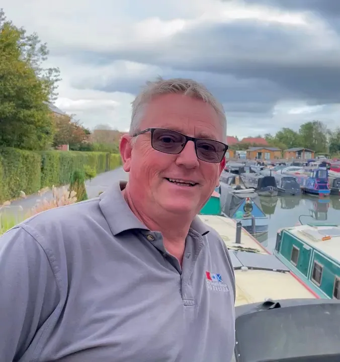 A smiling man wearing glasses and a grey polo shirt is standing in front of a marina with boats and greenery in the background.