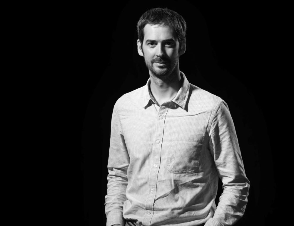 A man with short hair and a beard, wearing a light button-up shirt, is standing against a plain dark background.