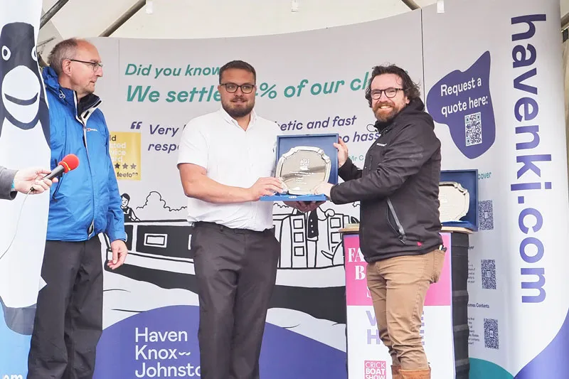 Three men participate in an award presentation, holding two plaques for the Crick Favourite Boat in Show, standing in front of banners promoting a business.