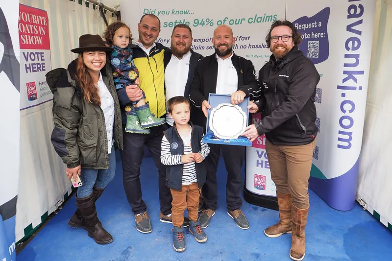 A group of six people, including two children, stand indoors next to promotional banners. One person proudly holds an award plaque for the 