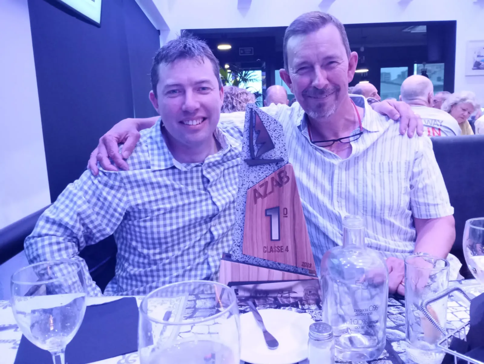 Two men sit together at a dinner table, smiling and holding a triangular wooden trophy labeled 