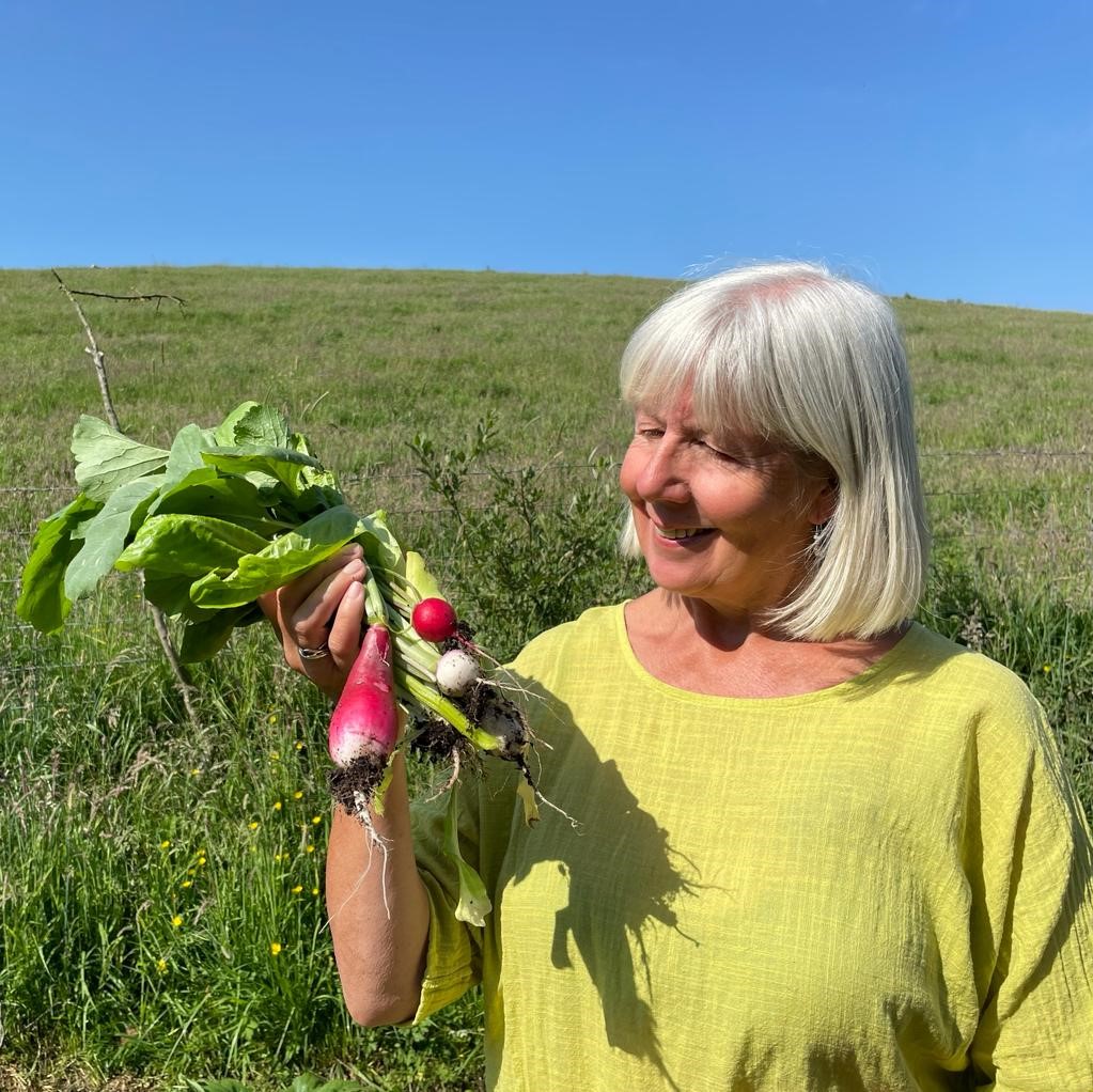 A woman in a yellow shirt holds freshly harvested radishes while standing outdoors in a grassy field under a clear blue sky.