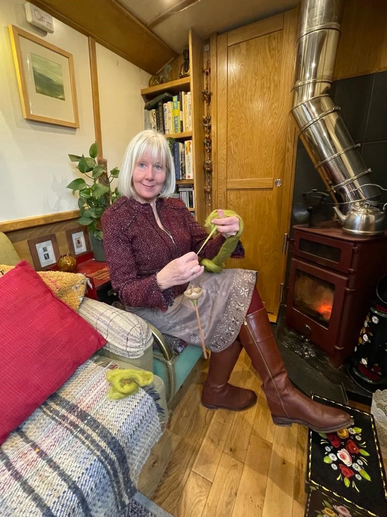 A woman sits in a cozy room, spinning wool by a wood-burning stove. She smiles at the camera, surrounded by books and plants.