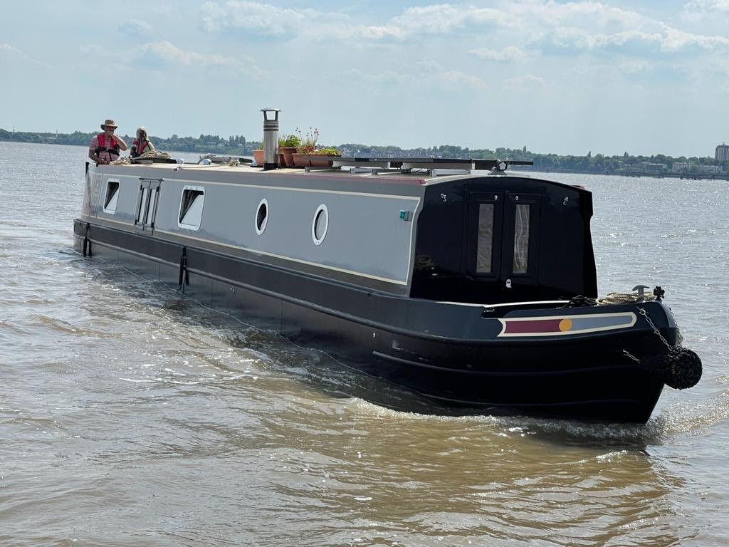 A narrowboat cruises on a body of water with two people at the helm under a partly cloudy sky.