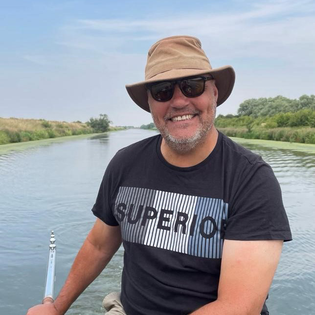 A smiling person wearing sunglasses and a hat sits in a boat on a calm river, with greenery on both sides under a blue sky.