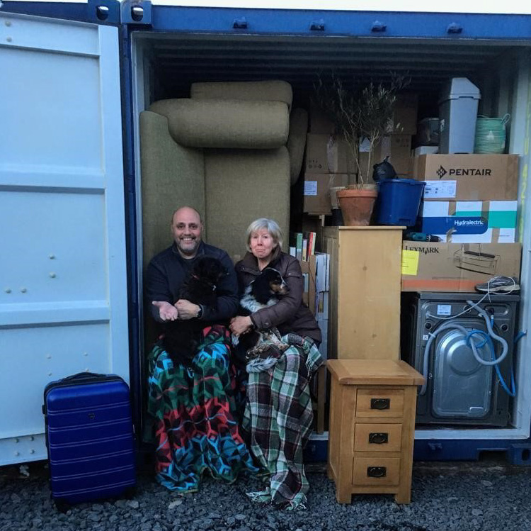 Two people sit with pets in a storage container filled with furniture, boxes, and household items, with a blue suitcase outside.