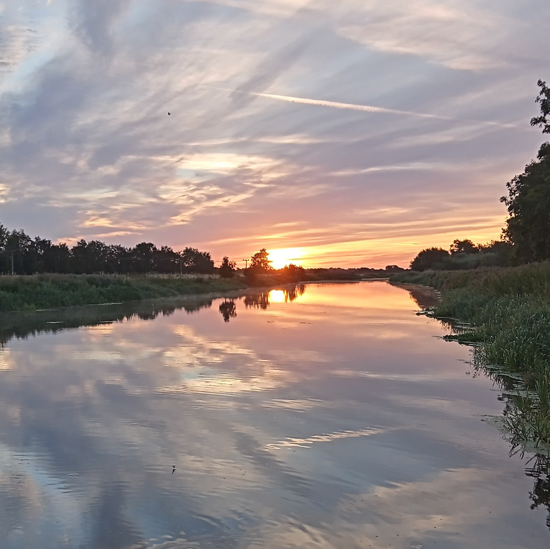 A sunset reflecting on a calm river, with trees lining the banks and a sky streaked with clouds.