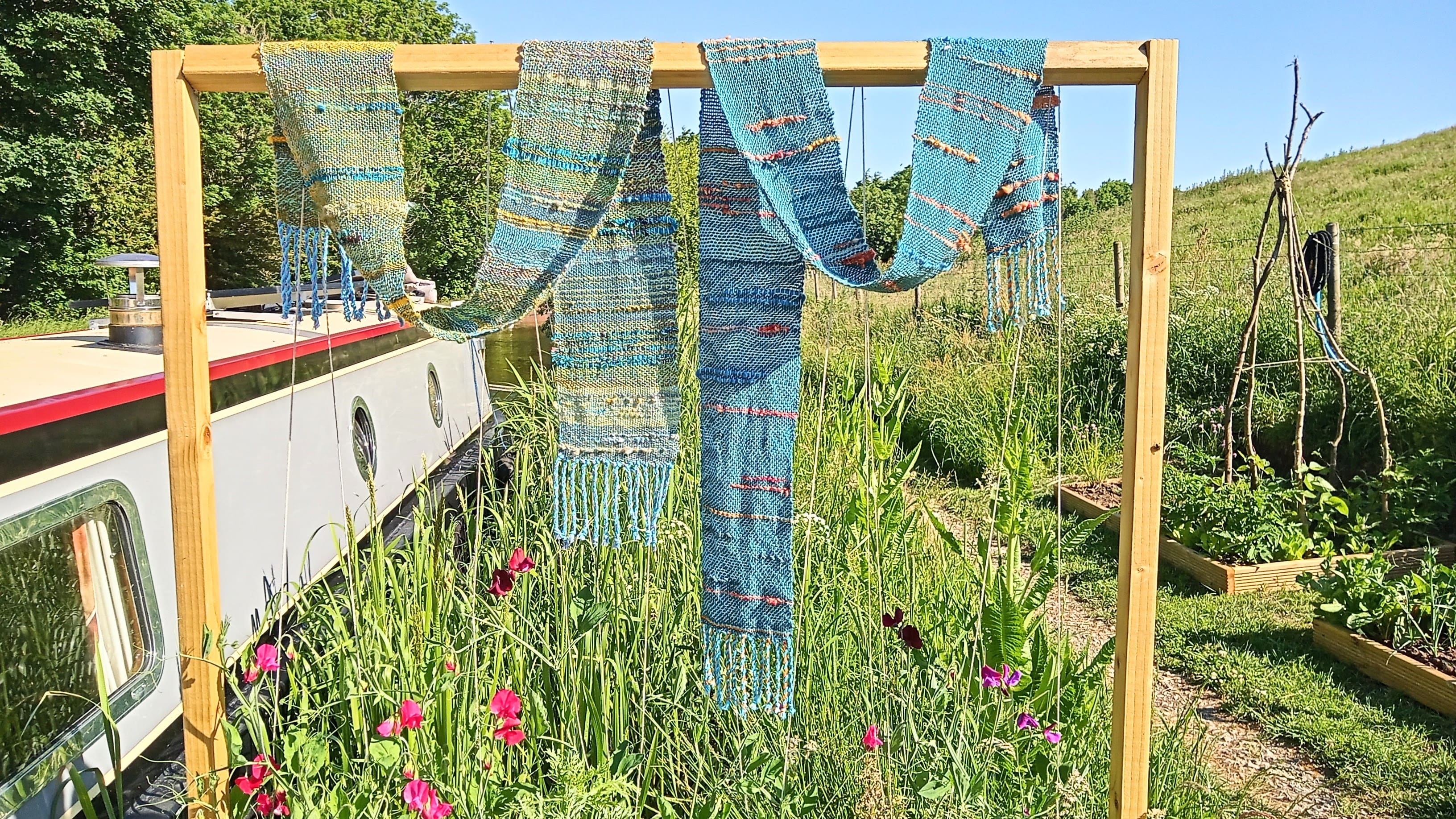 Three blue woven scarves hanging on a wooden frame outdoors, with a narrowboat and garden in the background on a sunny day.