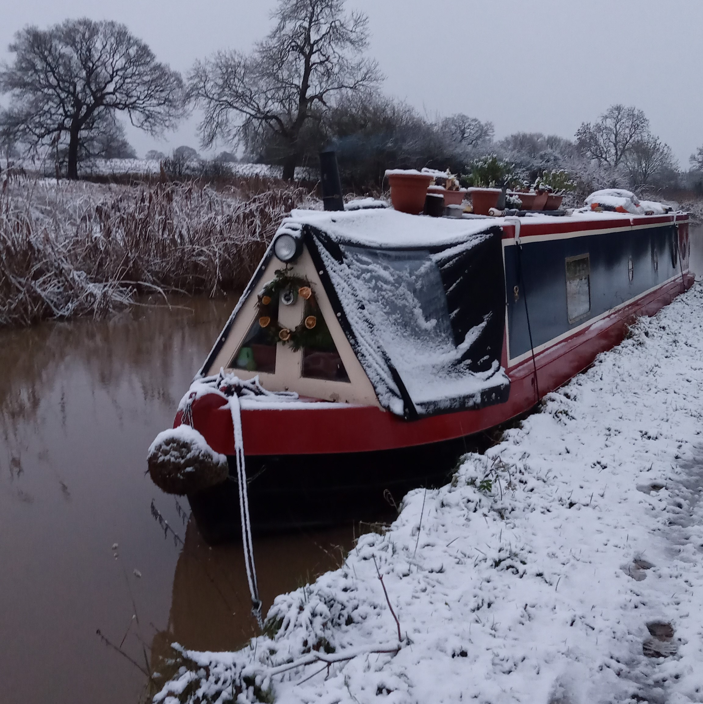 A canal boat covered in snow moored by the side of a canal, with trees and a snowy landscape in the background.