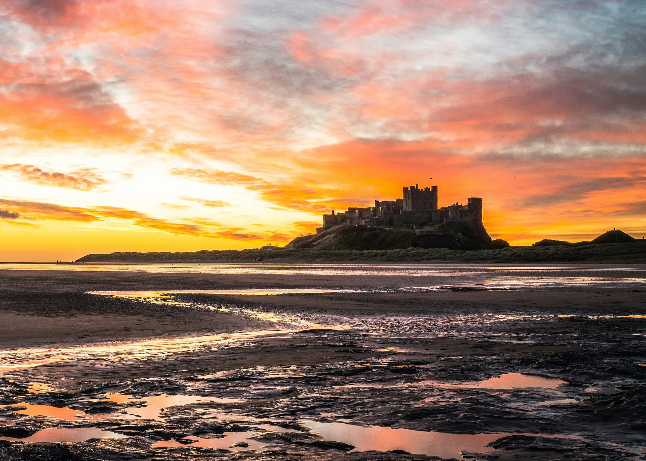 Distant castle silhouetted against vibrant orange and pink sunset sky, with dark, wet sand and shallow water pools in the foreground.