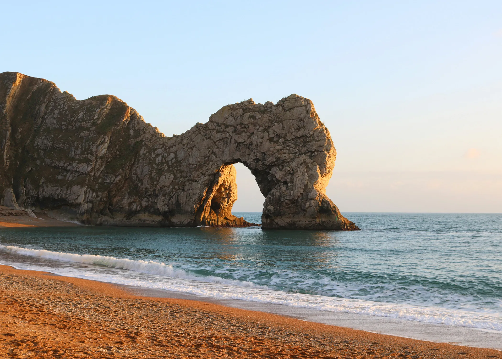 A natural limestone arch known as Durdle Door rises from the sea along a sandy beach under a clear sky at sunset.