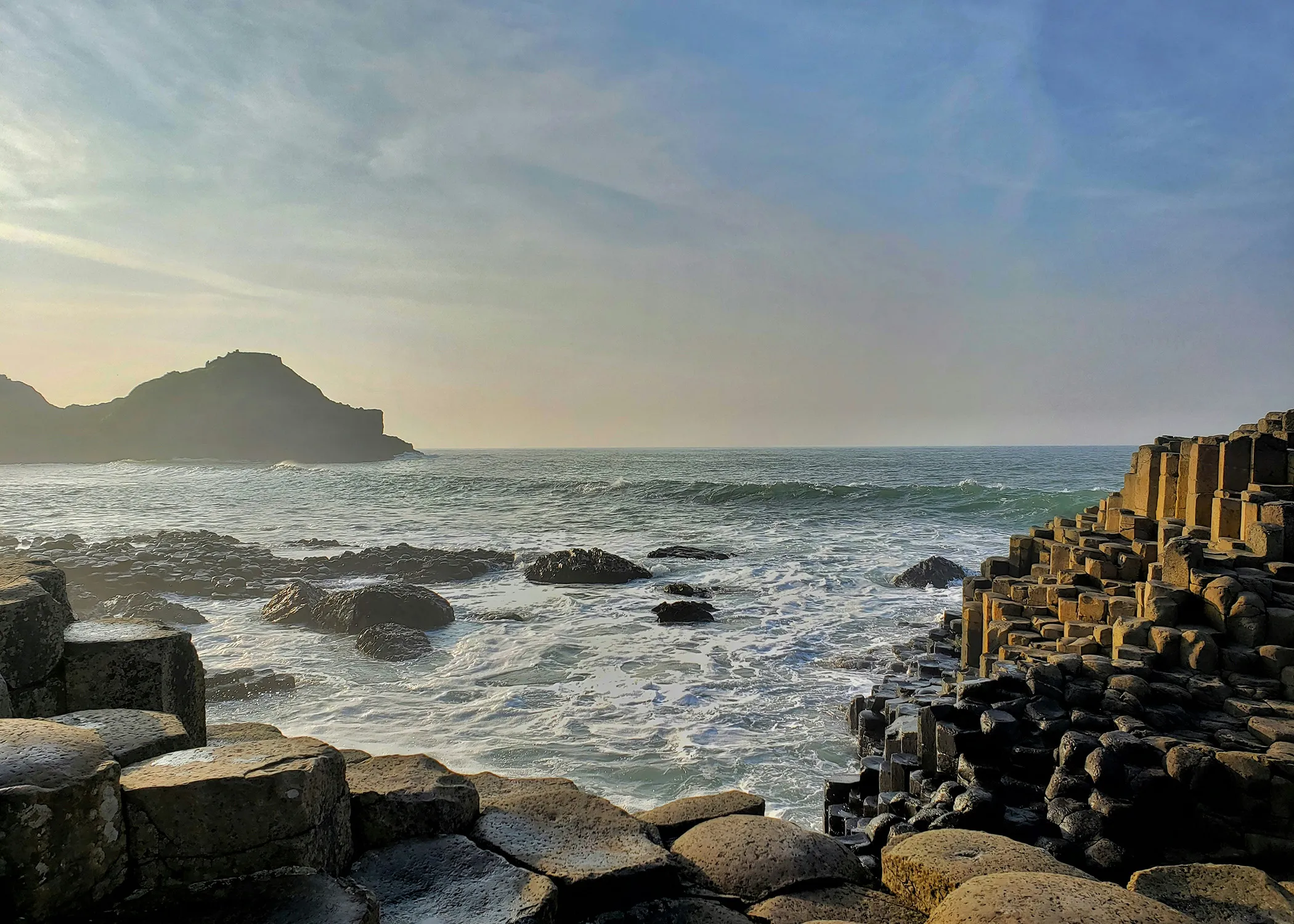 View of the sea meeting the rocky coastline with hexagonal basalt columns in the foreground and a hilly silhouette in the background.