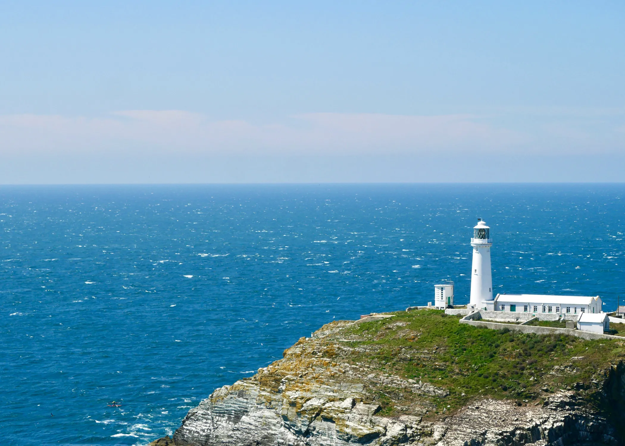 A white lighthouse stands atop a rugged cliff overlooking a vast, calm ocean under a clear blue sky.