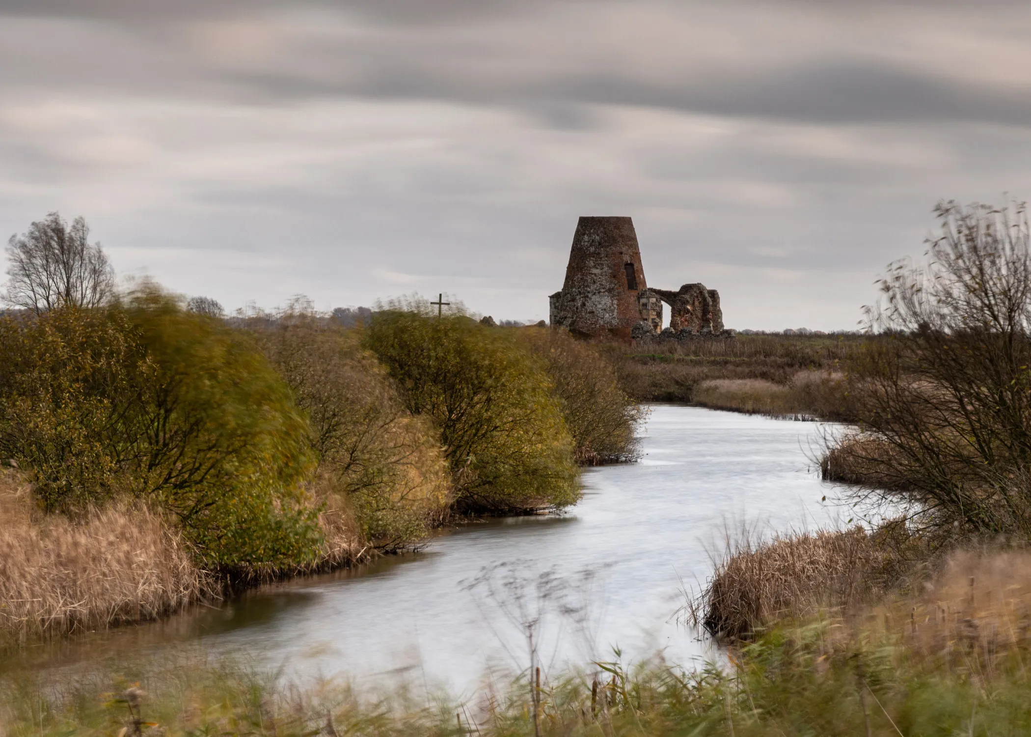 A tranquil river flows through marshland, with an old, partially ruined brick building in the distance under a cloudy sky.