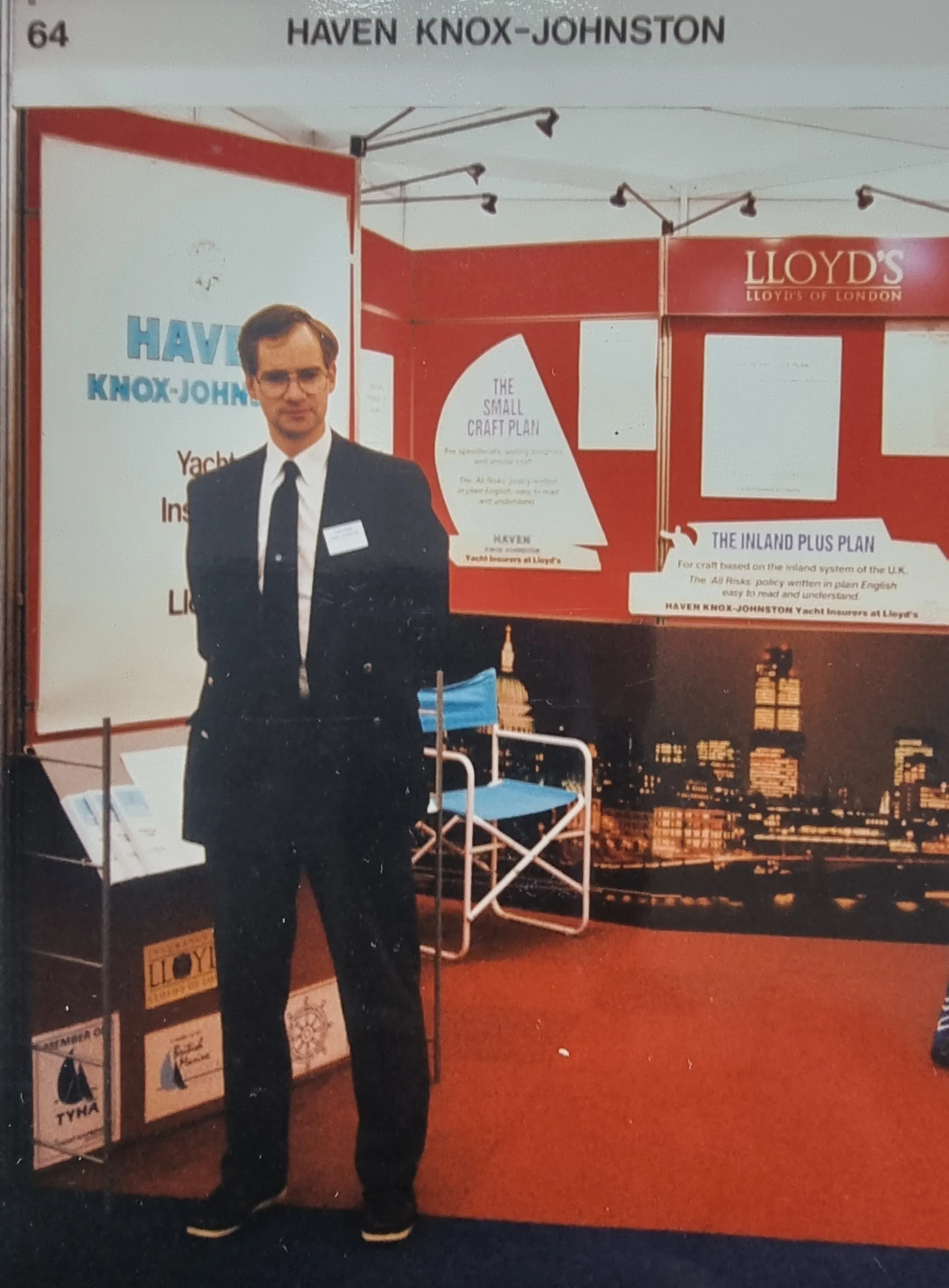 A man in formal attire stands in a trade show booth for Haven Knox-Johnston and Lloyd's, with promotional materials behind him.