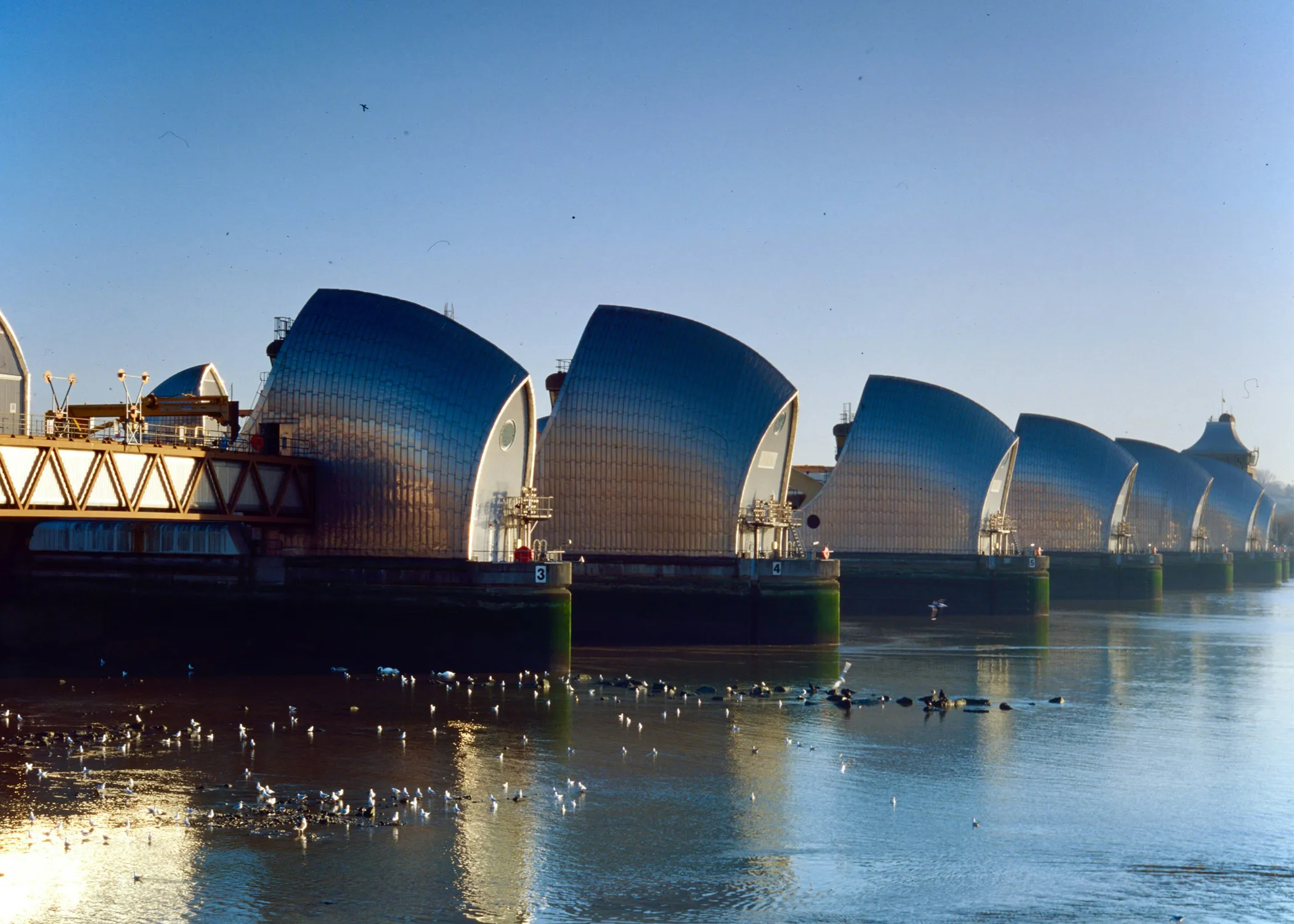 The image shows the Thames Barrier, a series of large silver flood barriers across the River Thames under a clear blue sky.