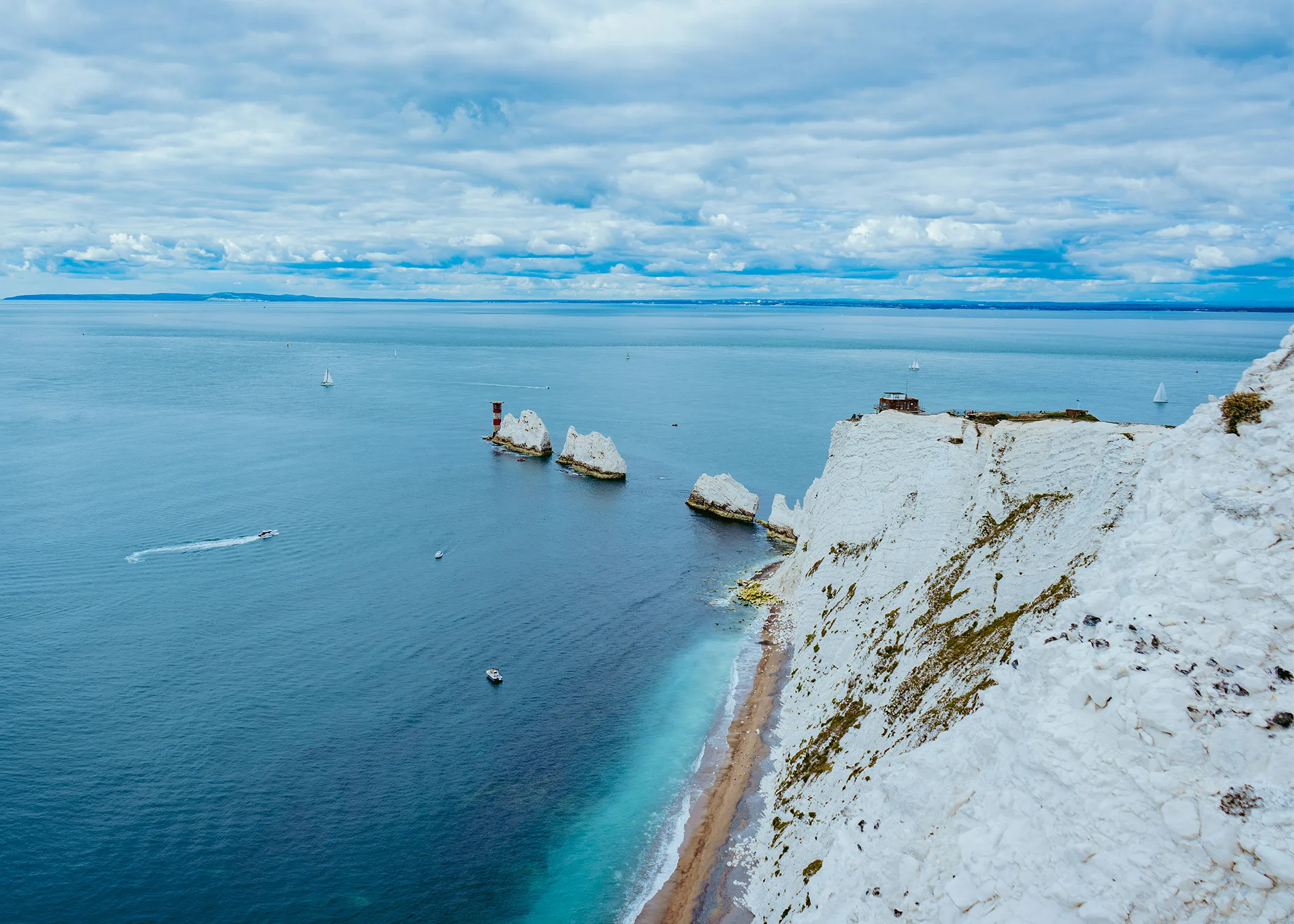 Cliffs overlooking a blue ocean with a few boats and yachts visible in the water, under a partly cloudy sky.