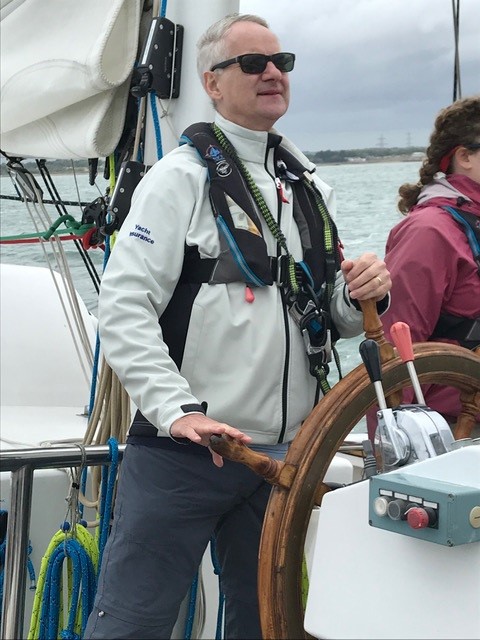 A man wearing a white jacket and sunglasses steers a sailboat, while another person stands nearby.