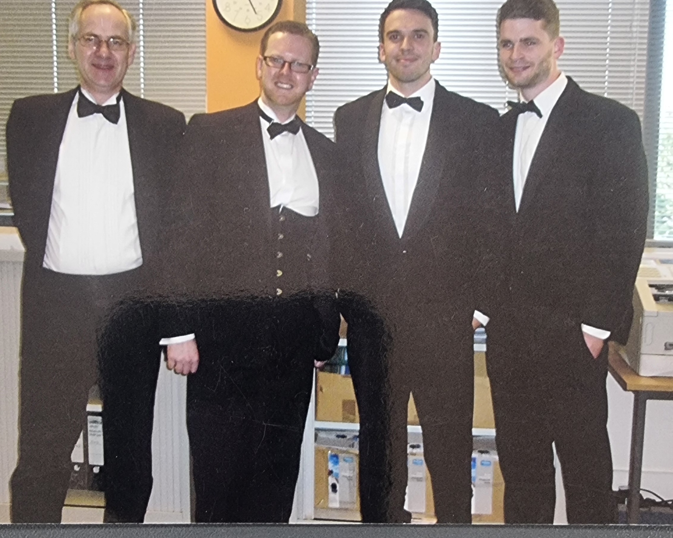 Four men in formal black-tie attire standing side by side in an office setting with blinds and a clock in the background.