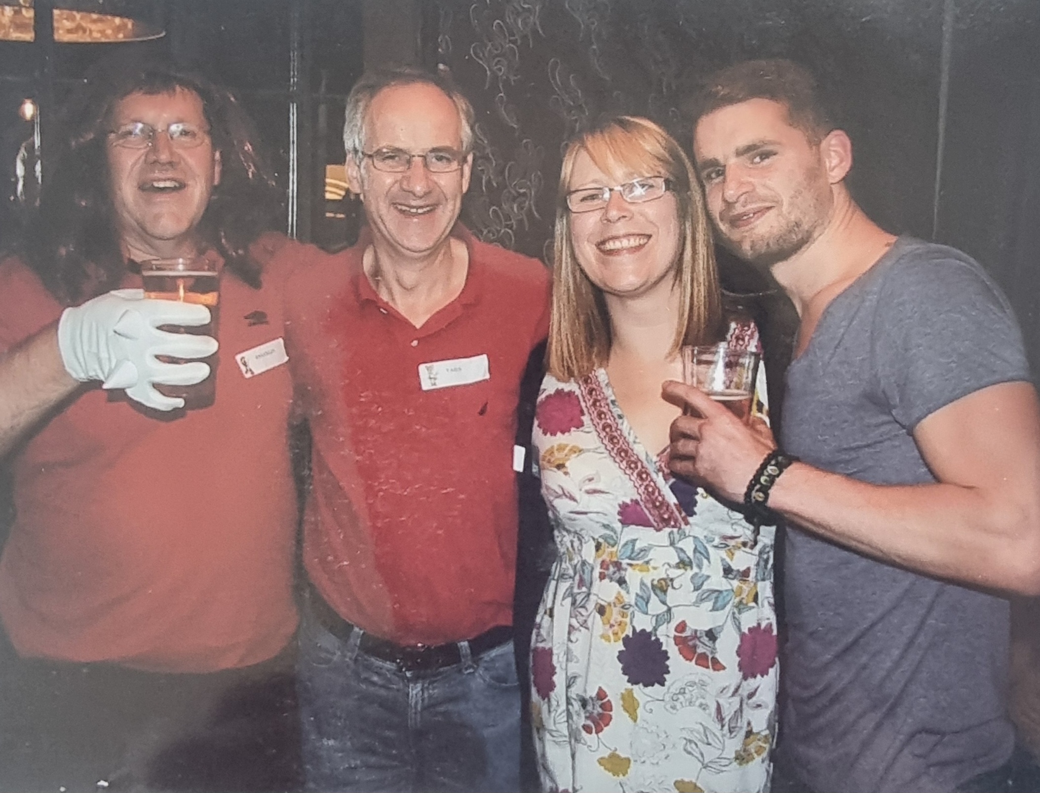 Four people are standing together, smiling at the camera while holding drinks. They are dressed casually and wearing name tags.