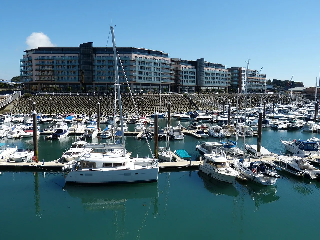 A marina filled with various boats and yachts docked, with a large modern building in the background under a clear blue sky.