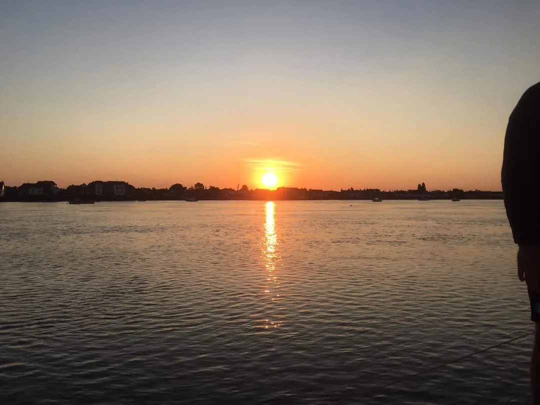 Sunset over a calm body of water with a clear sky, silhouetted shorelines in the distance, and a person partially visible on the right.