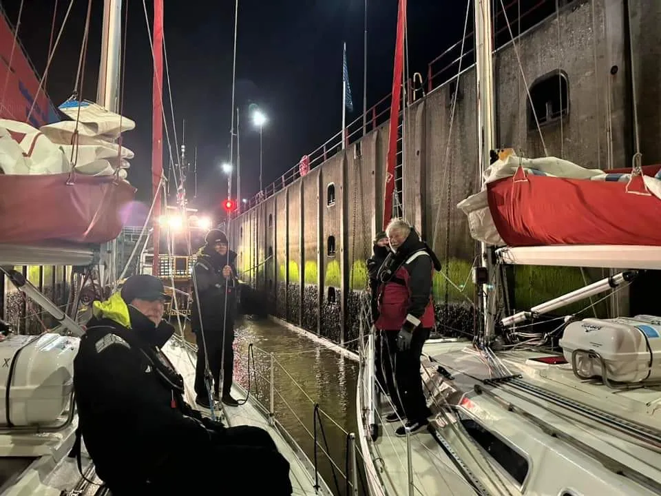 Three people in sailing gear stand on yachts in a lock at night, attending to ropes and waiting for water to rise.