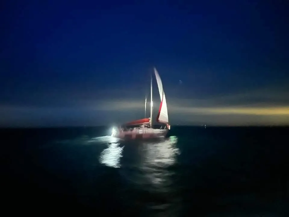 A sailboat with red and white sails is illuminated on dark, calm waters under a night sky with a faint horizon.