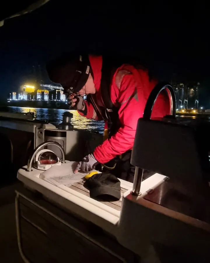 A person in a red jacket uses a flashlight to read documents on a boat at night, with illuminated buildings in the background.