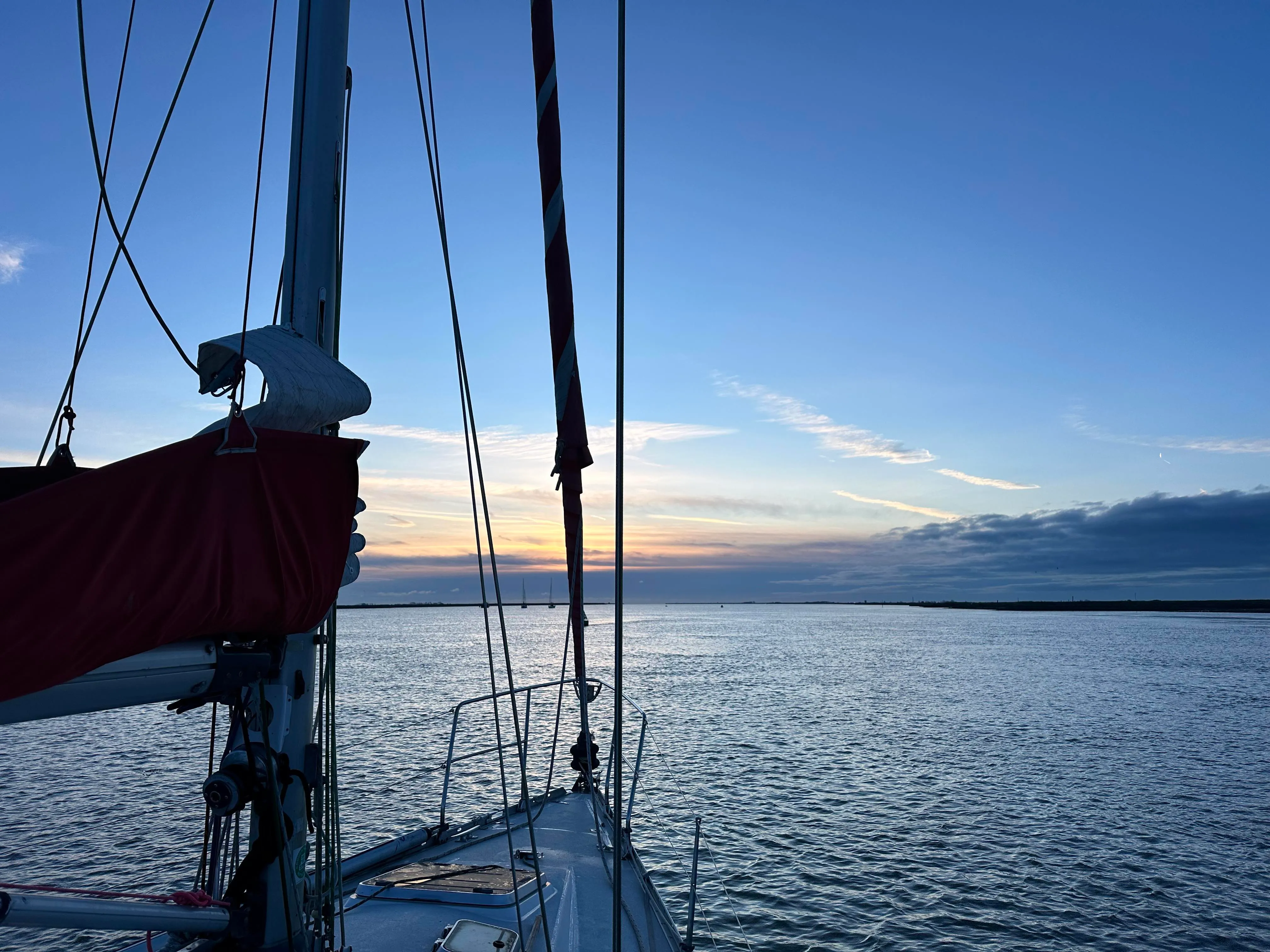 A calm evening view from the bow of a sailboat on the water, with a sunset over the horizon and a clear sky.