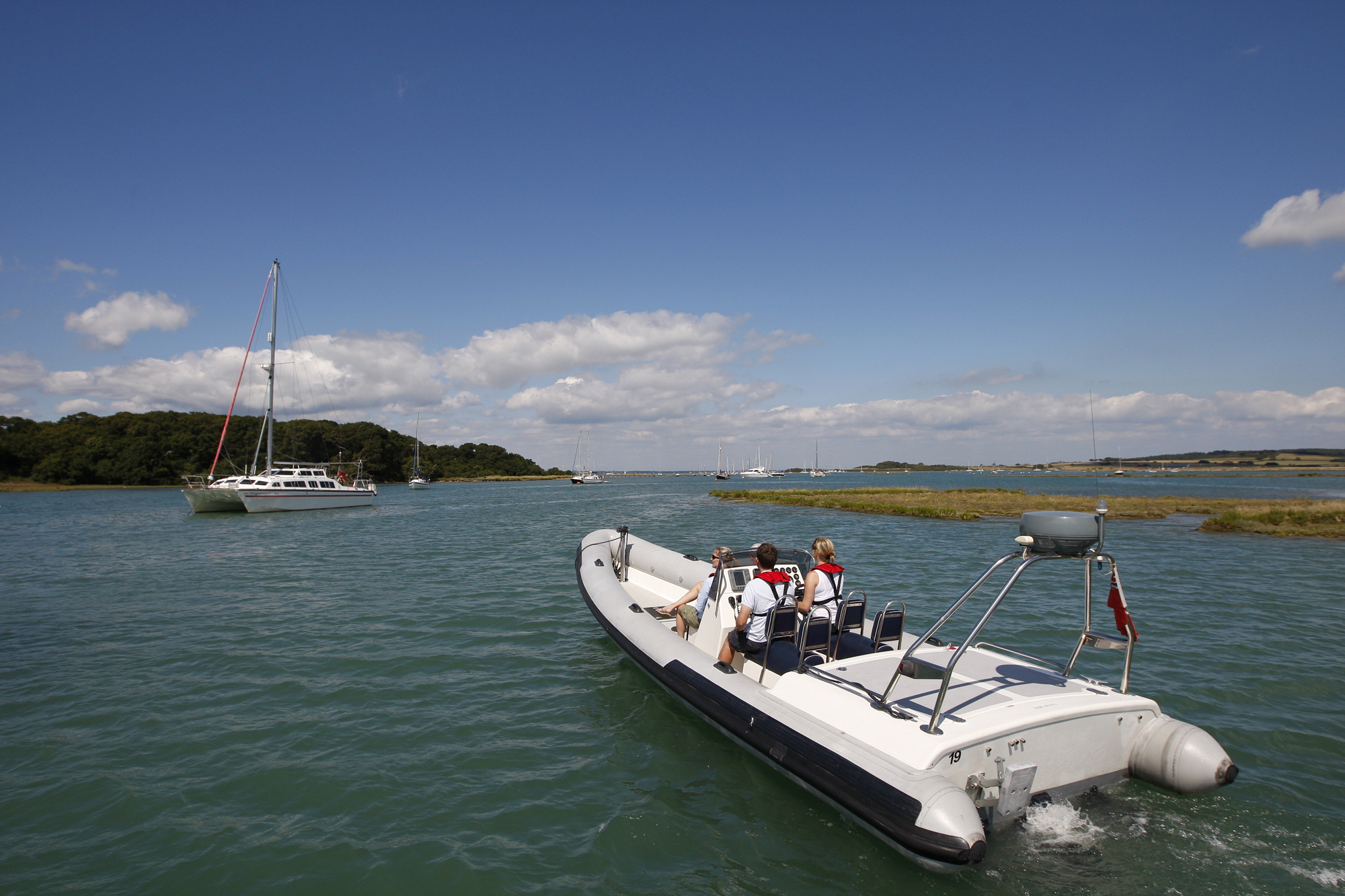 A small boat with four people navigates a waterway on a sunny day, with a sailboat visible in the background against a wooded shore.