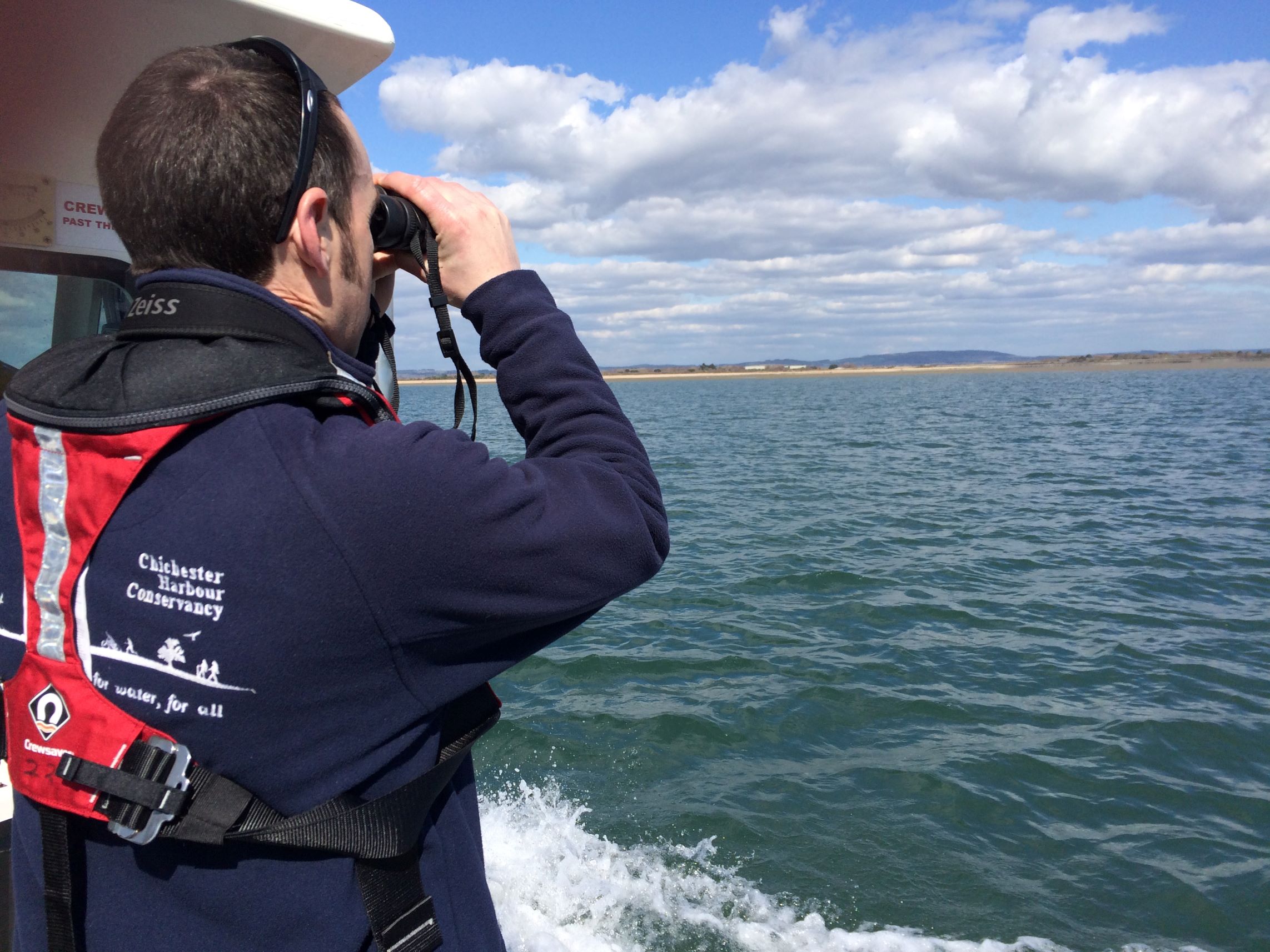 Person in a red life jacket using binoculars on a boat looking at the horizon over water under a partly cloudy sky.