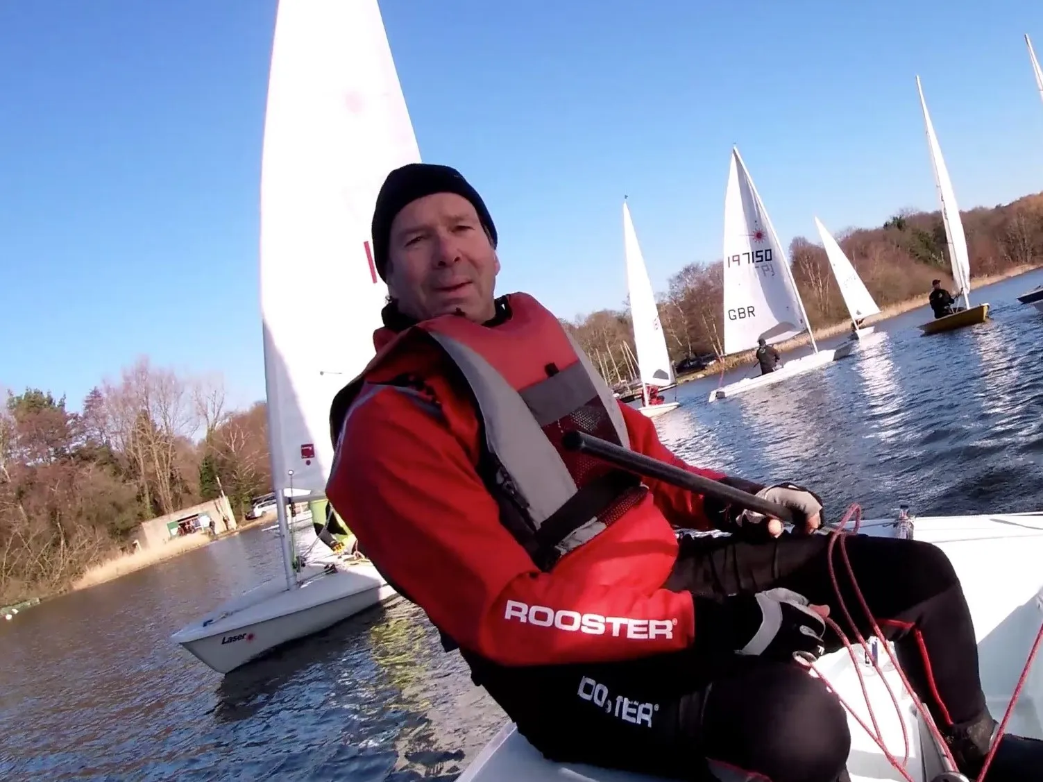 A man in a red sailing suit is sitting in a small sailboat on a lake, surrounded by other sailboats on a clear day.