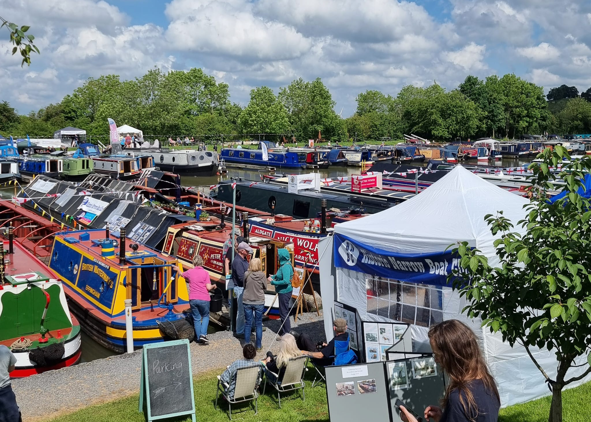 People attending a canal boat festival, with numerous boats docked and various stalls set up under a partly cloudy sky.